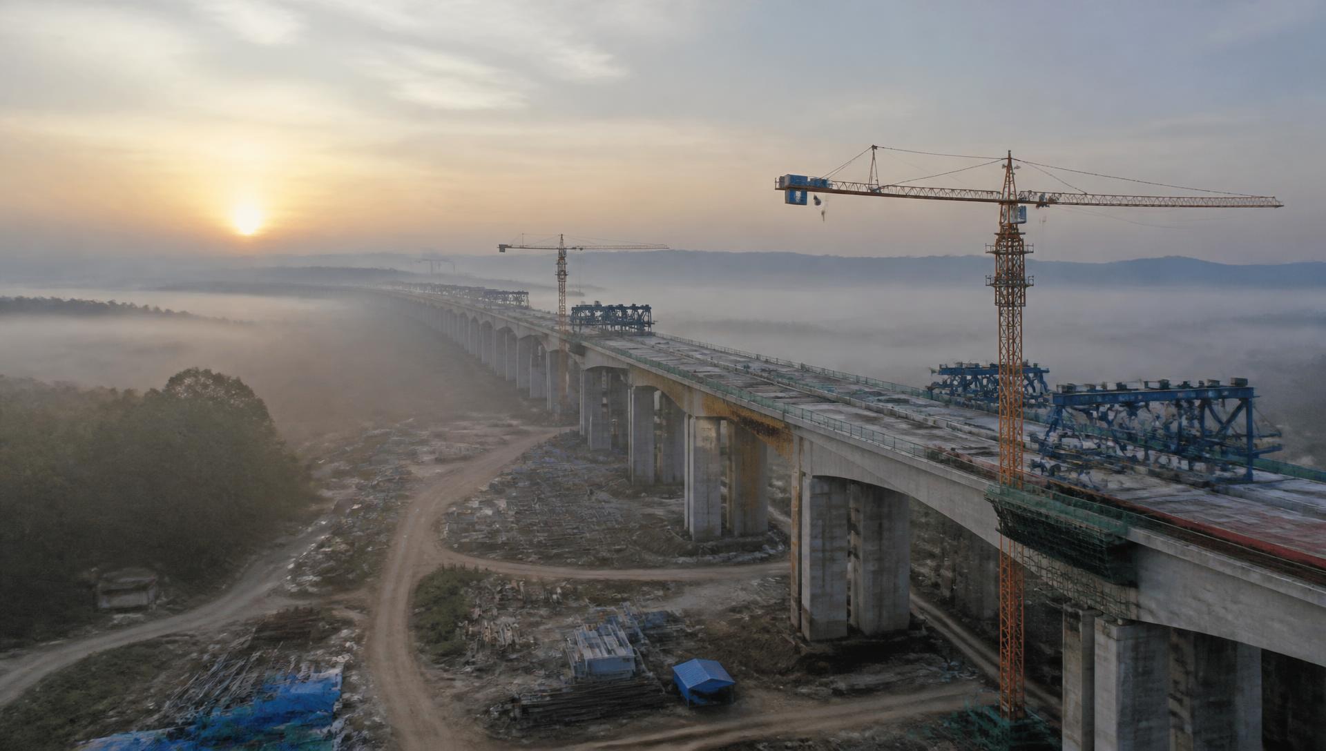 Aerial view of a viaduct under construction at dawn, tower cranes rising above valley mist